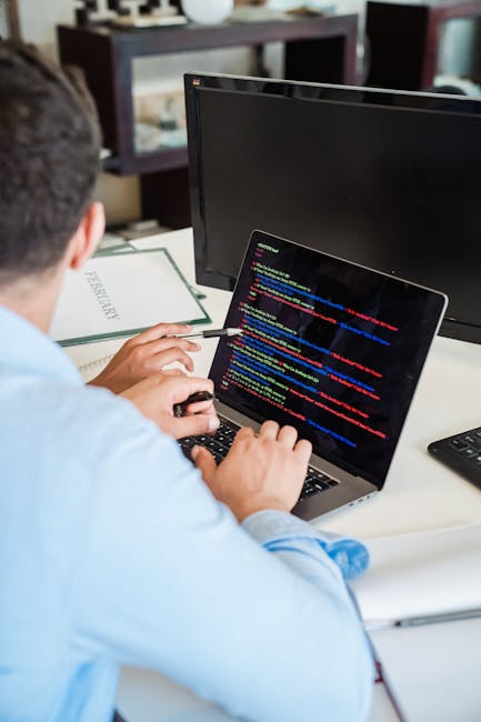 A person working at a desk with a laptop displaying lines of colorful code, positioned in front of a large external monitor. The individual, seen from behind, is holding a mouse with their right hand